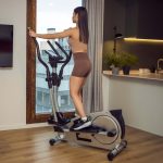 Woman in athletic wear exercising on an elliptical trainer in a home gym area by a large window.