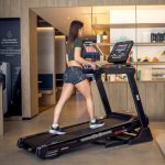 Woman runs on a black treadmill in a modern gym area with wooden shelves and a cafe-like background.