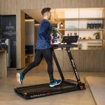 Man in a blue jacket runs on a treadmill in a modern gym with wooden shelving in the background.