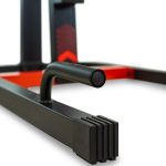 Close-up of a black metal exercise machine frame with a rounded grip bar and stacked rectangular pads; red accents are in the background.