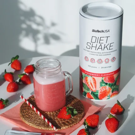Pink strawberry diet shake in a mason jar on a woven coaster, with a BioTechUSA Diet Shake canister and scattered strawberries on a white table.