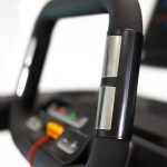 Close-up of a treadmill handrail with a black grip and silver bands, featuring a red safety key on the console in the blurred background.