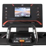 Treadmill console with a central touchscreen showing a desert road, surrounded by program and fitness icons; foreground handlebars.