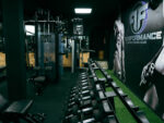 Gym interior with rows of dumbbells on a rack and weight machines against a dark wall with 'Performance' branding on the right.