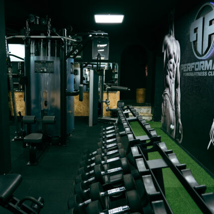 Gym interior with rows of dumbbells on a rack and weight machines against a dark wall with 'Performance' branding on the right.