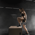 Female athlete in a black sports outfit stepping onto a wooden plyometric box while holding a weight plate in one hand amidst gym fog.
