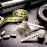 Fitness scene with a measuring tape, powder in a scoop, capsules, and a weight plate on a gym mat surface in low lighting.