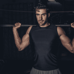 Muscular man in a sleeveless shirt lifting a barbell across his shoulders in a rack, looking focused in a dim gym.