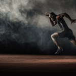 Male sprinter in a black racing suit mid-sprint on a red track, with dramatic smoke in the background.