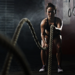 Athletic woman in a sports bra and shorts performing battle rope exercises in a dim gym.