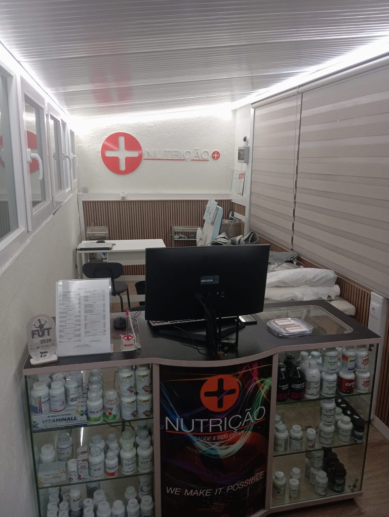 Counter with a computer and glass display case filled with vitamin bottles in a nutrition shop; red cross logo and 'NUTRIÇÃO' on the back wall