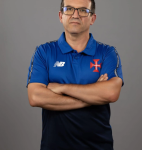 Man with arms crossed, wearing a blue polo with a crest on the chest and New Balance logo, neutral gray background, glasses.