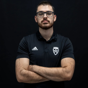 Portrait of a man with glasses and a beard, arms crossed, wearing a black polo with an Adidas logo and a shield crest.