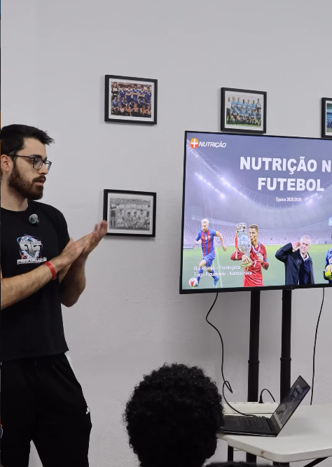 Presenter in black shirt with glasses claps beside a screen showing a football nutrition presentation; laptop on a table and an audience member visible.