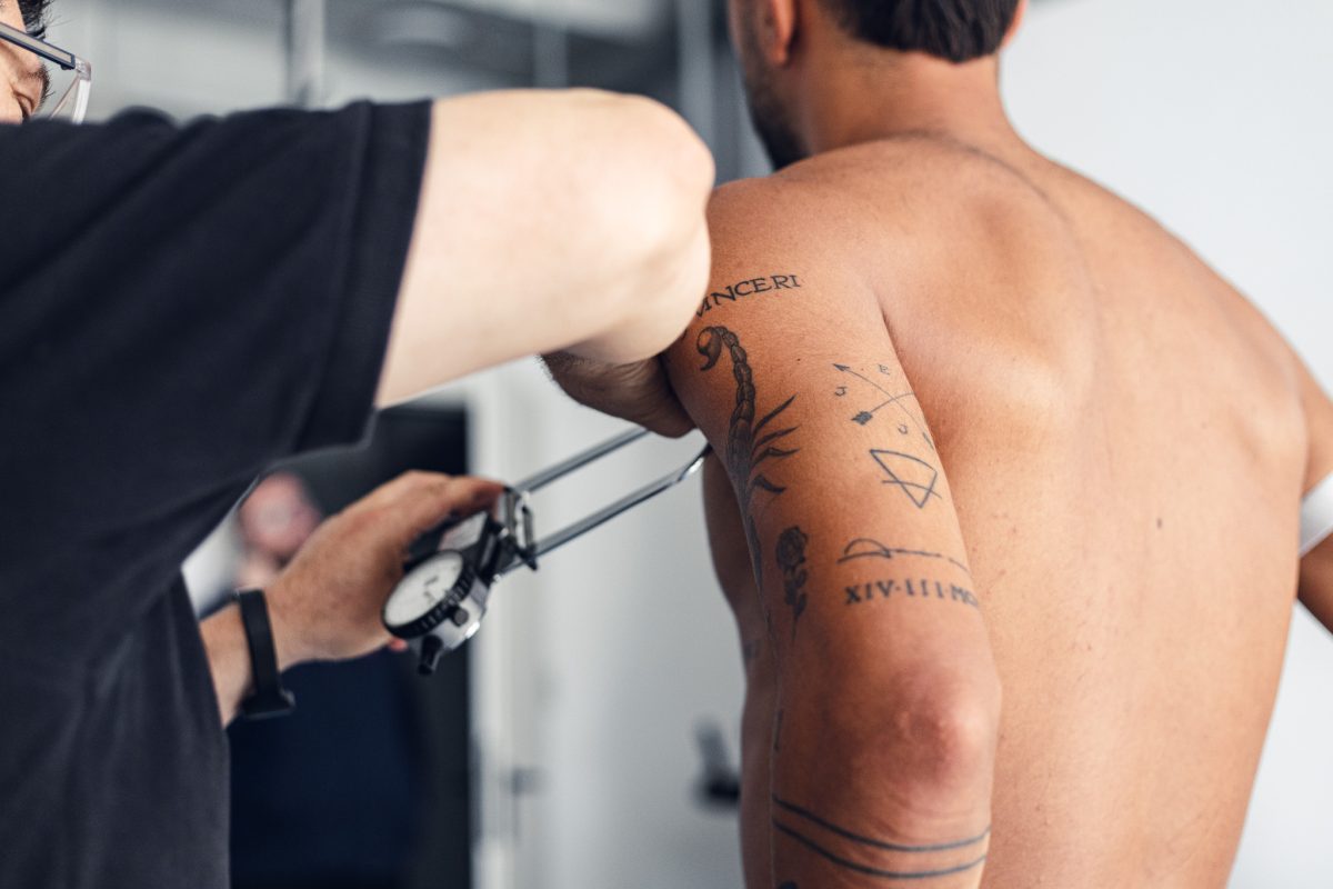 Tattoo artist in a black shirt using a tattoo machine on a shirtless man's upper arm, showing tattoos on the arm.