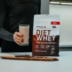 Bag of PhD DJET WHEY Lean Protein Powder Belgian Chocolate beside a glass of chocolate shake and chocolate pieces on a white kitchen counter, person in black shirt nearby.