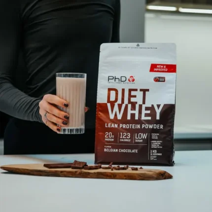 Bag of PhD DJET WHEY Lean Protein Powder Belgian Chocolate beside a glass of chocolate shake and chocolate pieces on a white kitchen counter, person in black shirt nearby.