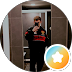 Person standing in a doorway in a dim hallway, wearing a red shirt; round orange badge with a star in the bottom-right corner.