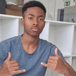 Young man in a blue T-shirt posing in a room with white shelves, making a double shaka gesture toward the camera.