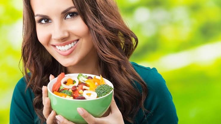 Woman smiling outdoors while holding a green bowl of colorful mixed salad with vegetables.