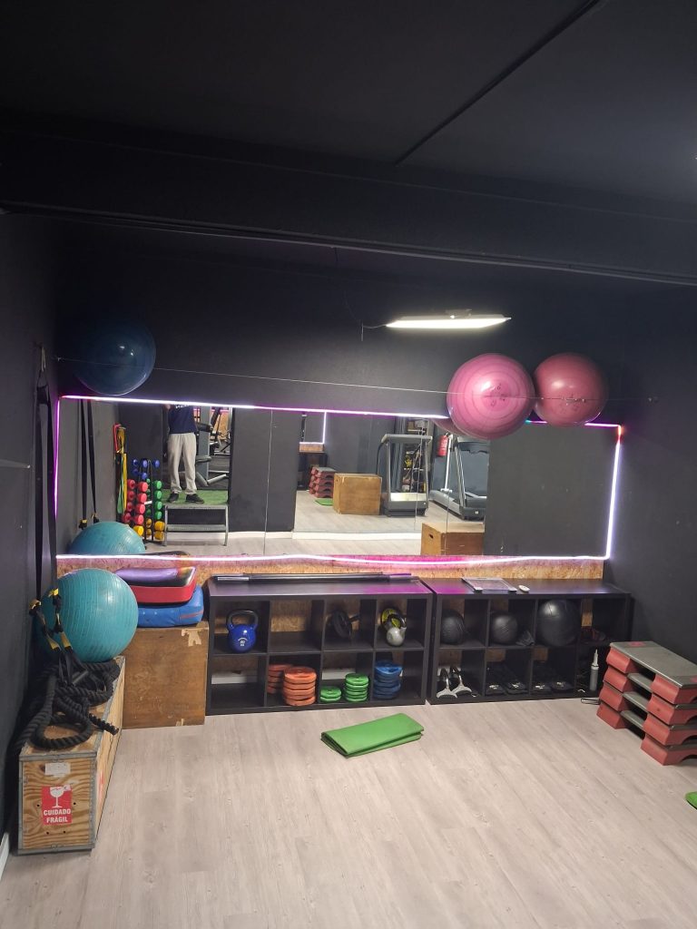 Gym studio with mirrored wall and pink exercise balls, kettlebells, weights, and mats arranged along shelving units.