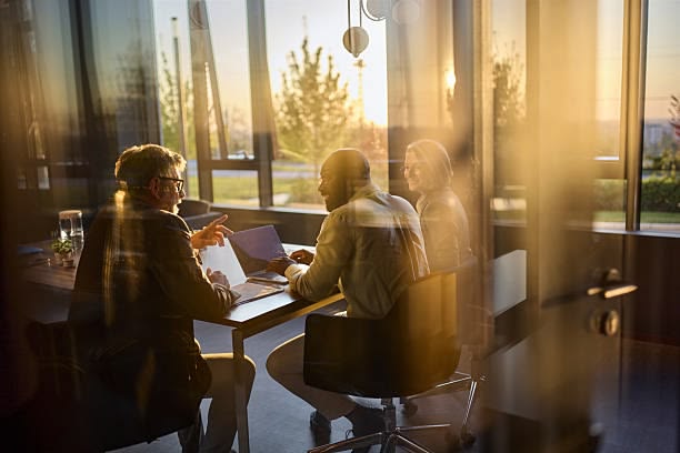Three colleagues discuss documents around a table in a sunlit glass-walled meeting room at sunset.