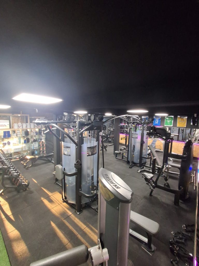 A dimly lit gym filled with resistance machines, free weights, and benches arranged in rows, reflecting in mirrors along the walls.