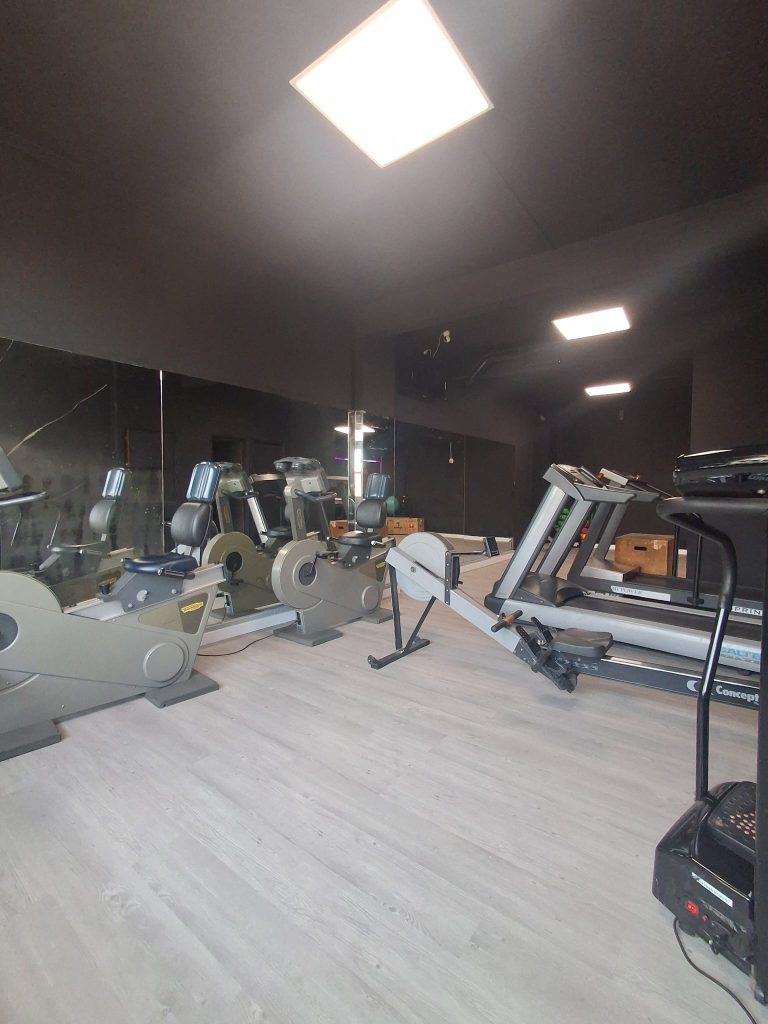 Glimmering cardio area of a dark gym with multiple treadmills and ellipticals along a mirrored wall under bright square ceiling lights