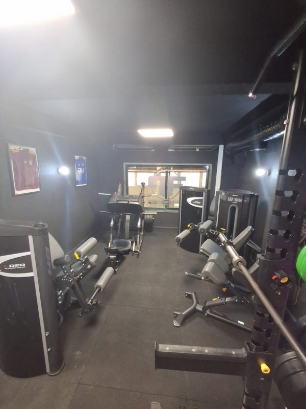 Compact gym interior with multiple weight machines, benches, and racks along dark walls; window at the far end.