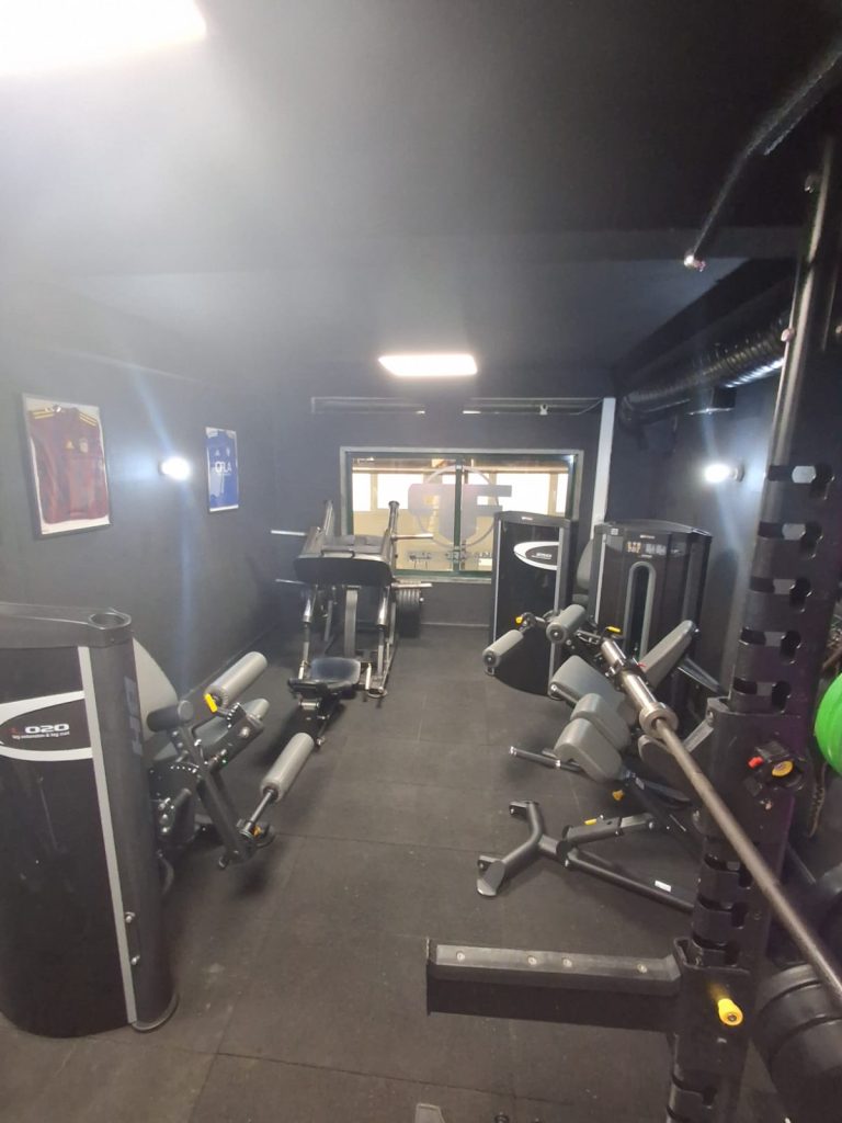 Compact gym interior with multiple weight machines, benches, and racks along dark walls; window at the far end.