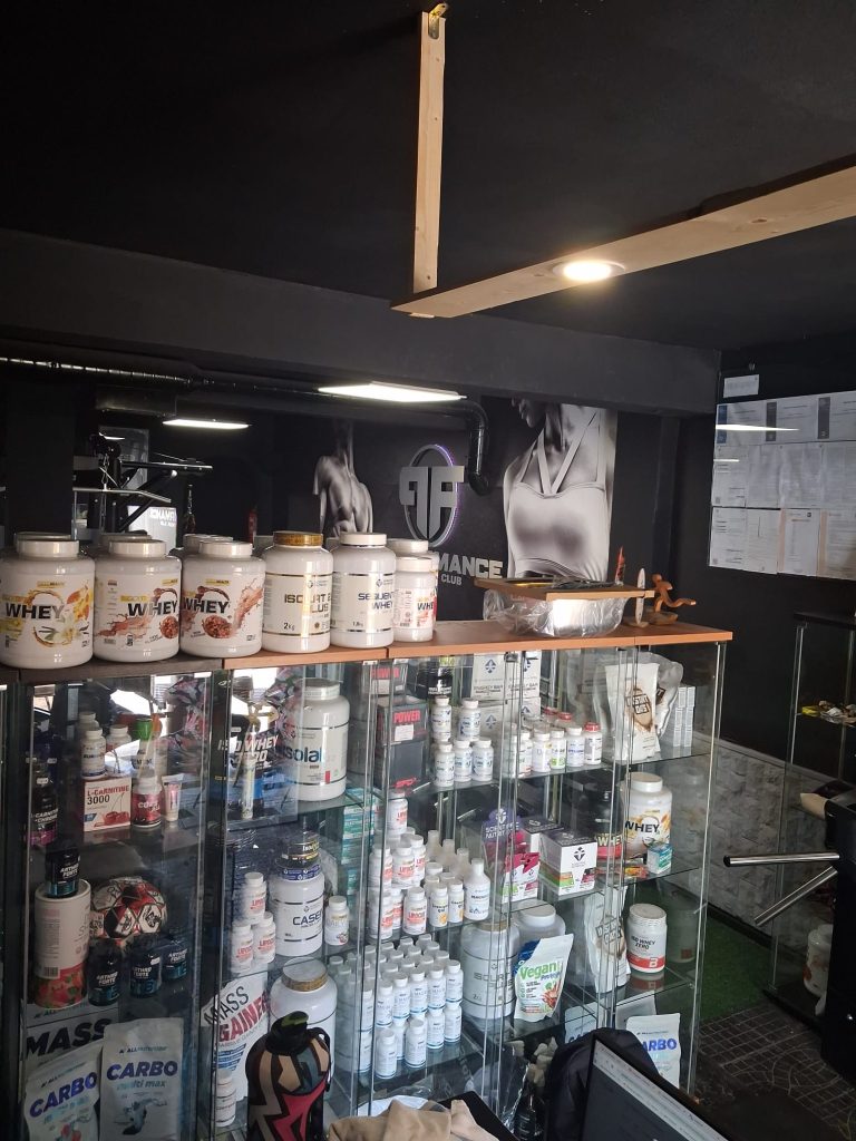 Inside a gym supplement shop with glass display cases full of protein powders and vitamins on shelves under black walls and posters.