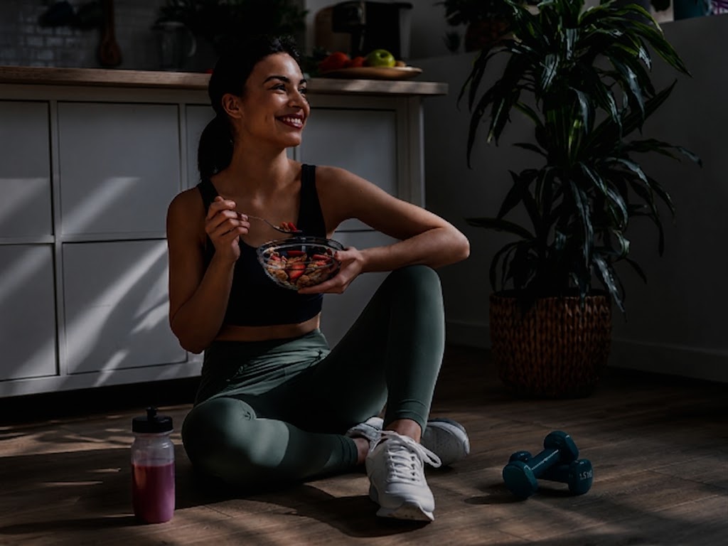 Woman in workout clothes sits on the floor eating fruit from a bowl, smiling in a home gym/kitchen setting with a pink smoothie bottle nearby and teal dumbbells on the floor.