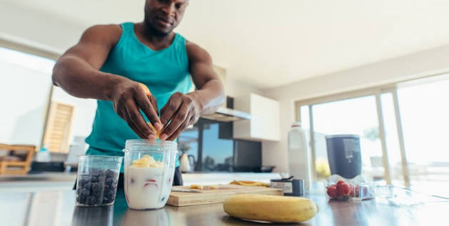 Man in a teal tank top sprinkles toppings into a yogurt cup on a bright kitchen counter.