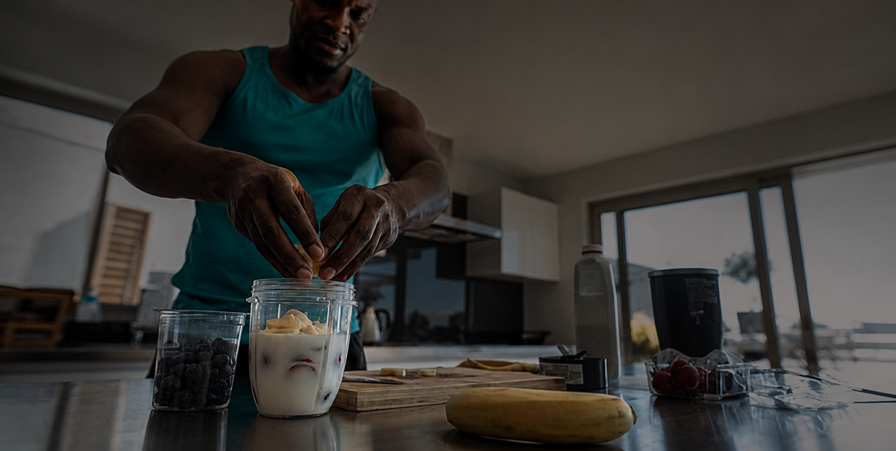 Man in a turquoise tank top adds banana slices to a yogurt parfait in a glass jar on a kitchen counter filled with berries and fruit.