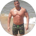 Man standing shirtless on a sunny beach, wearing green camouflage swim trunks.