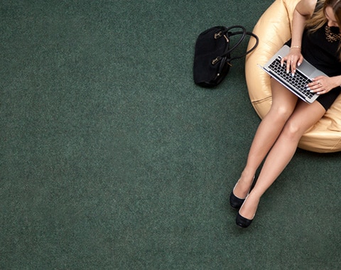 Woman in a black dress sits on a round beige chair on a green carpet, typing on a laptop with a black handbag beside her.