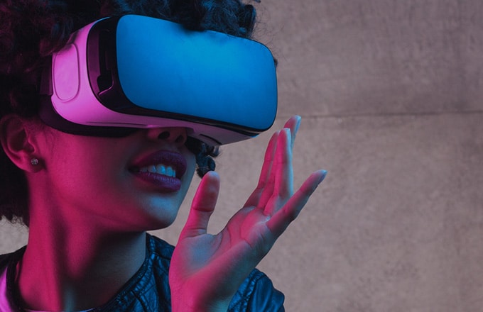 Person wearing a blue VR headset with pink lighting, reaching forward as if interacting with a virtual environment.
