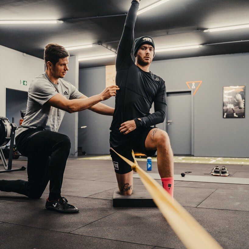 Trainer assists client performing a resistance-band stretch in a gym, client kneeling on a block with one arm raised.