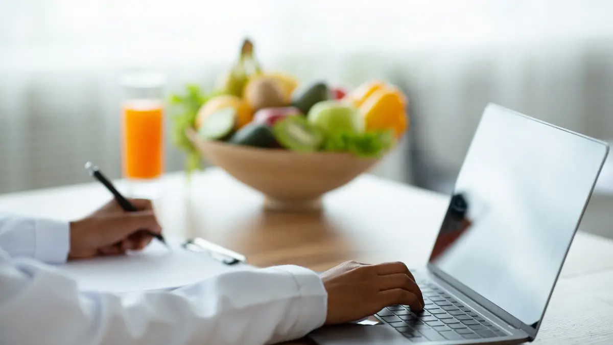 Person typing on a laptop at a desk with a bowl of fruit and a glass of juice in the blurred background, suggesting a healthy_WORK setup.