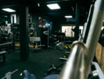 Wide-angle gym interior with rows of weight machines, benches, and cable stations in a dimly lit, dark-painted space with mirrors and posters on the walls