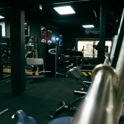 Wide-angle gym interior with rows of weight machines, benches, and cable stations in a dimly lit, dark-painted space with mirrors and posters on the walls
