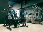 Row of stationary bikes in a dim gym with a dark green wall and wooden floor.