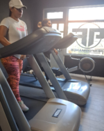Two women exercise on treadmills in a bright gym, one wearing a white Puma shirt, cap, and pink patterned leggings nearby the window.
